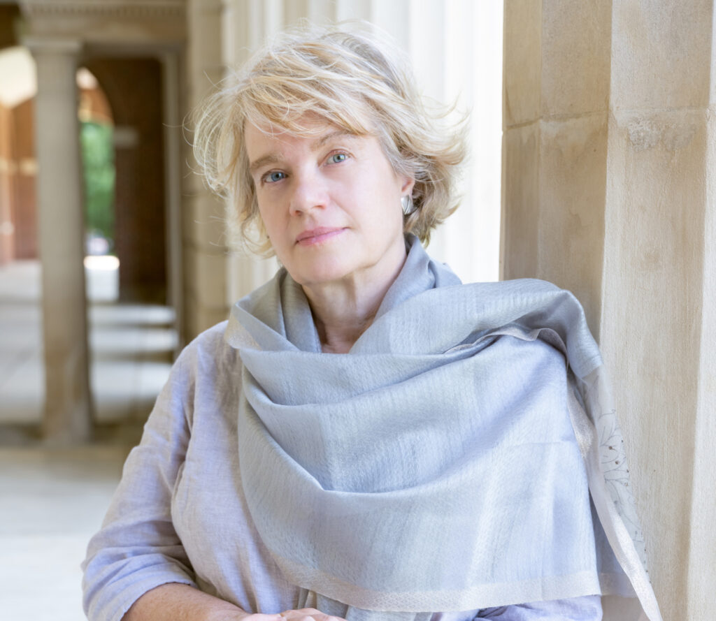 A portrait of the author and educator Melora Wolff. She is a white person, with blonde hair in a layered bob. She is wearing a lavender top and a light blue shawl. She is leaning against the wall of a marble building that may be on a college campus. There is a pillar in the background.